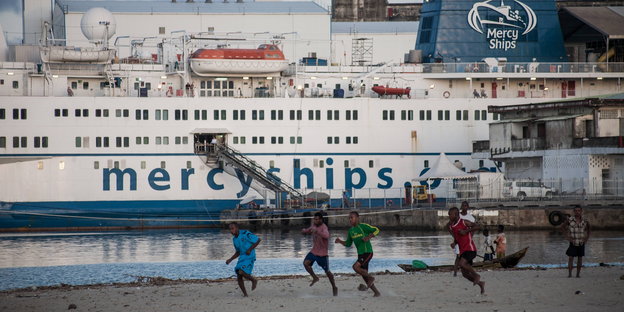 Ein Schiff im Hafen, am Strand spielen Menschen Fußball