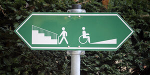 Ein Schild zeigt in zwei Richtungen, rechts ist eine Rampe für Rollstuhlfahrer, links eine Treppe für Fußgänger