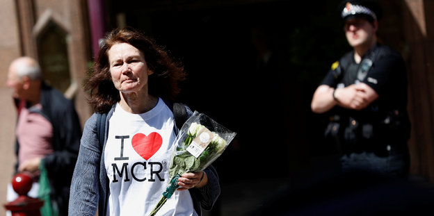Eine Frau geht an einem Polizisten vorbei, sie trägt weiße Blumen und ein weißes T-Shirt, auf dem I love MCR steht