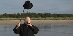 Brian Cox als Winston Churchill am Strand, eine Zigarre im Mund und seinen Bowler mit seinem Gehstock über dem Kopf haltend