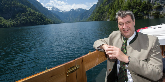 Markus Söder lehnt an der Reling eines Bootes und lächelt, im Hintergrund See und Berge