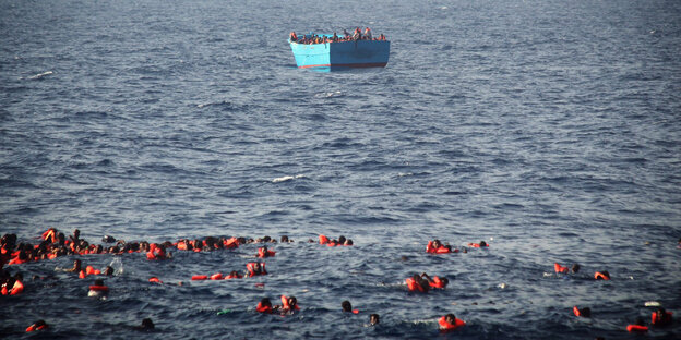 Szene auf offenem Meer, im Hintergrund ein blaues Holzboot, im Vordergrund viele Menschen in Schwimmwesten im Wasser