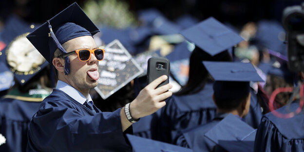 Ein Universitäts-Absolvent in Talar trägt eine Sonnenbrille, streckt die Zunge raus und macht eine Selfie mit seinem Handy