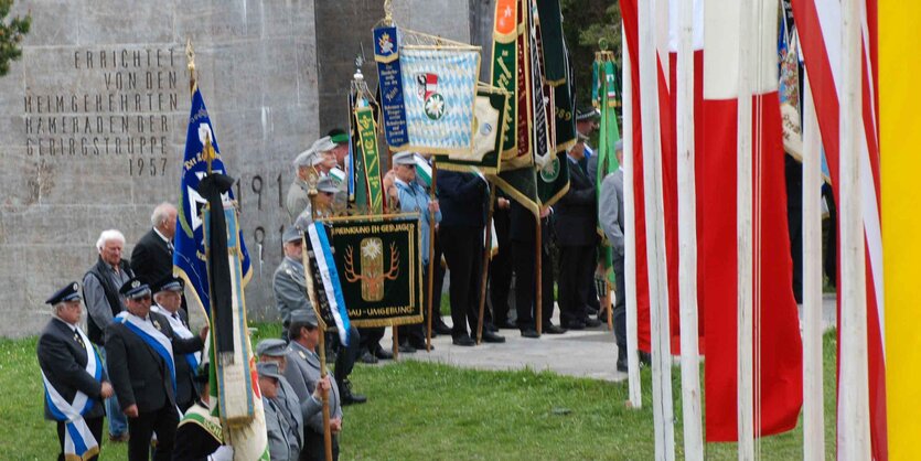 Auf einer grünen Wiese ist im Hintergrund ein großer Felsen, im Vordergrund mehrere Menschen in Uniformen mit Bannern und Flaggen