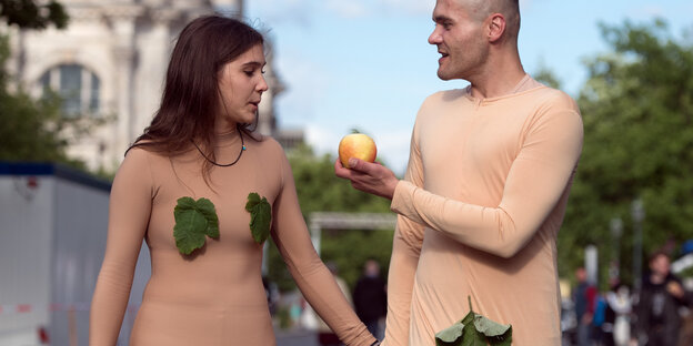 Eine Frau und ein Mann haben sich als Adam und Eva verkleidet. Er hält einen Apfel in der Hand. Sie schaut auf den Apfel. Sie halten Händchen.