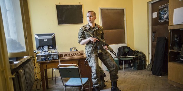 Ein uniformierter Mann mit Sturmgewehr lehnt an einem Schreibtisch