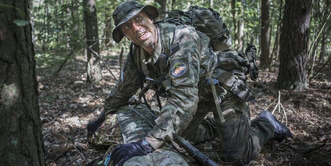 Ein Mann in Flecktarn kniet in einem Wald