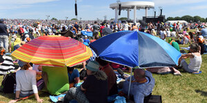 Viele Menschen sitzen auf einem weiten Feld in der Sonne, zwei von ihnen schützen sich mit bunten Regenschirmen