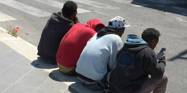 Vier junge Flüchtlinge sitzen nebeneinander auf einer Bordsteinkante, die Rücken gekrümmt. Einer spielt mit seinem Handy