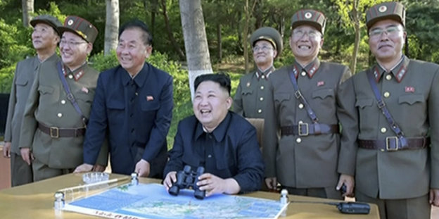 Nordkoreas Diktator Kim Jong Un sitzt zwischen Generälen und lacht wie ein Kind, in den Händen hält er ein Fernglas