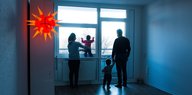Eine Familie steht in der Dämmerung am Fenster ihrer unbeleuchteten Wohnung und schaut in die Ferne