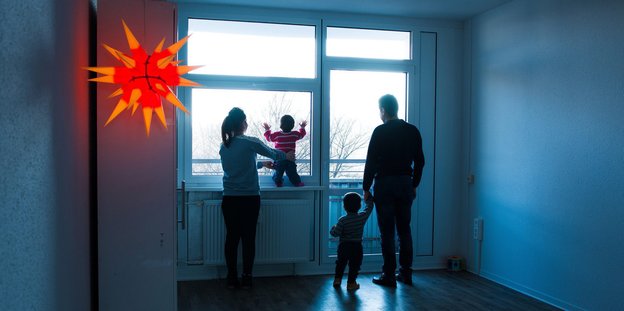 Eine Familie steht in der Dämmerung am Fenster ihrer unbeleuchteten Wohnung und schaut in die Ferne