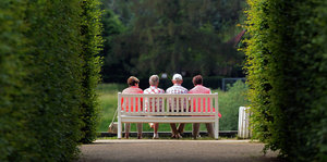 Vier ältere Menschen sitzen auf einer weißen Bank, gesäumt von zwei Hecken, und gucken ins Grüne