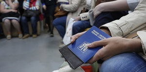 In einem Warteraum sitzende Menschen, im Vordergrund eine hand mit einem blauen Pass