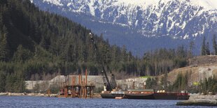 Seeufer am Fuß von Bergen, im Wasser ein Schiff und eine im Bau befindliche Plattform