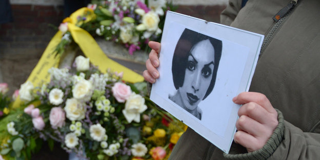 Hände einer Frau halten ein Foto der ermordeten Hatun Sürücü, im Hintergrund türmen sich Blumen