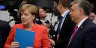 Merkel, in rotem Jackett, hält eine blaue Mappe fest in ihren Händen und guckt nach links, rechts von ihr steht Orban und guckt zu ihr