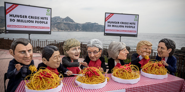 An einem Tisch sitzen Aktivisten, die Masken von Staatschef_innen tragen und Spaghetti essen, dahinter Schilder, auf denen „Hunger crisis“ steht