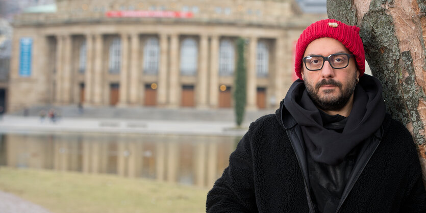 Mann mit roter Mütze, Bart und Brille lehnt an einem Baum