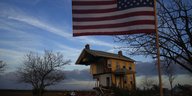 Ein zerstörtes Haus im Hintergrund vor einem dämmernden Himmel, im Vordergrund eine wehenede US-Flagge