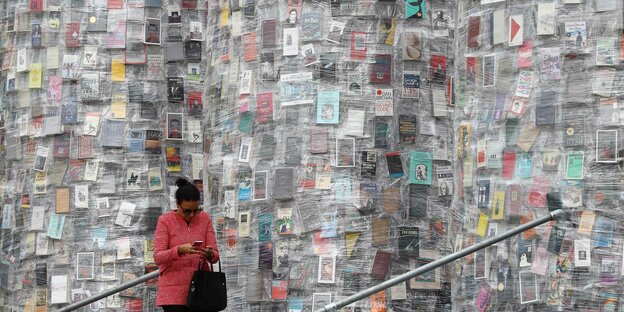Eine Frau in roter Jacke guckt auf das Smartphone in ihren Händen, hinter ihr das risige Kunstwerk "Tempel der Bücher"