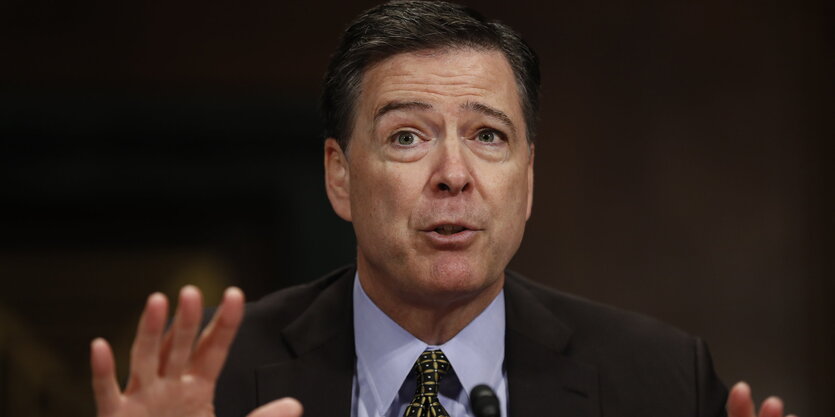 James Comey sitzt vor schwarzem Hintergrund, runzelt die Stirn und hebt beschwichtigend beide Hände