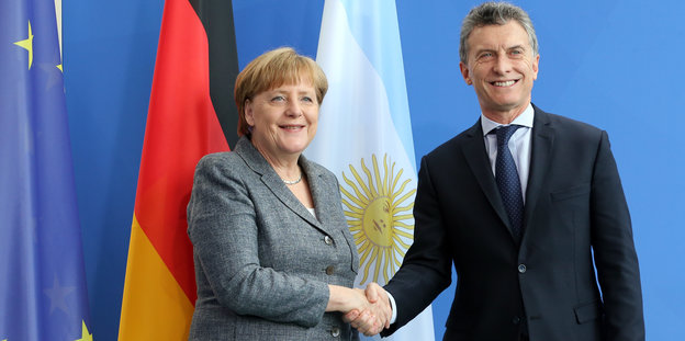 Ein Mann und eine Frau schütteln sich die Hände, im Hintergrund, die europäische und die argentinische Flagge