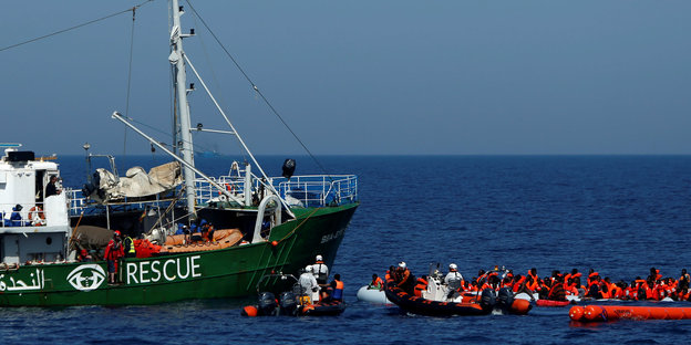 Ein Rettungsschiff mit der Aufschrift "Rescue" nimmt Geflüchtete in orangenen Rettungswesten auf, die vor dem Schiff in Rettungsbooten warten