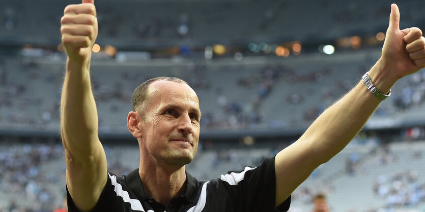 Ein Mann mit Glatze steht in einem Stadion und streckt seine Daumen in die Höhe