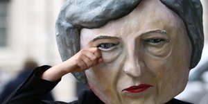 Eine Hand fässt an eine Pappmaske. Sie zeigt das Gesicht von Theresa May in Übergröße