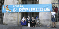 Frauen mit Kopftüchern sitzen unter einem Banner, das für die 6. Republik wirbt