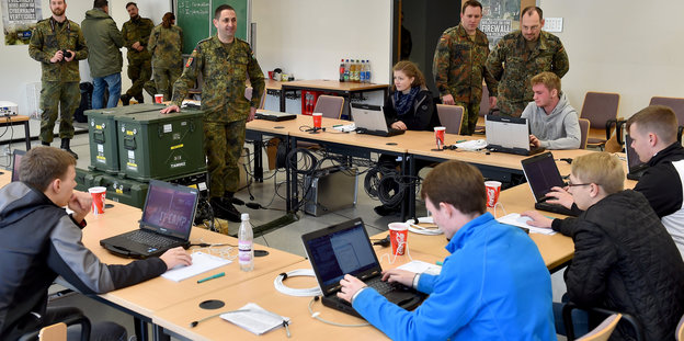 Ein paar Jugendliche sitzen mit Laptops an einem Tisch, davor stehen Menschen in Bundeswehr-Uniform