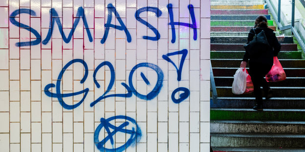 An einer Wand steht: „Smash G20“, im Hintergrund läuft eine Frau eine Treppe hoch