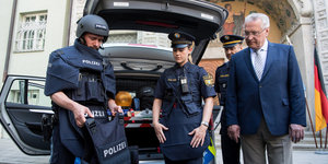 zwei Polizist_innen in Uniform vor einem Auto zeigen einem weißhaarigen Mann in Anzug schusssichere Westen