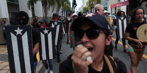 Eine Frau spricht in einen Lautsprecher, DemonstrantInnen tragen Fahnen