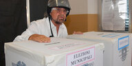 Ein Mann in weißem Hemd und mit Motorradhelm steht vor großen Wahlurnen aus Pappe