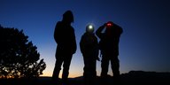 Drei Personen stehen im schwindenden Licht auf einem Hügel und blicken durch Nachtsichtgeräte