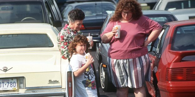 Zwei Frauen und ein Kind stehen zwischen Autos und trinken etwas
