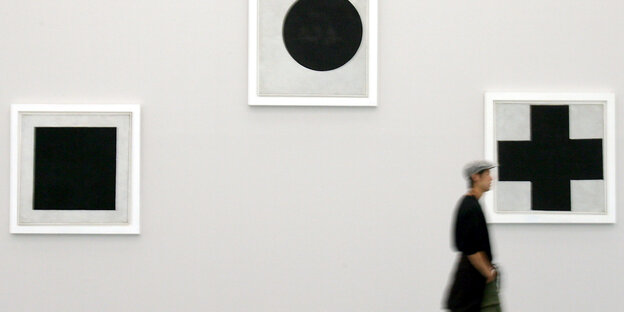 Eine Person läuft an einer Wand in einer Galerie vorbei, an der drei Bilder von Malewitsch hängen: ein schwarzes Quadrat, ein schwarzer Kreis und ein schwarzes Kreuz.