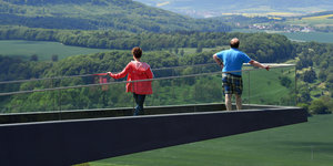 Zwei Menschen auf einer Aussichtsplattform, im Hintergrund eine grüne Landschaft