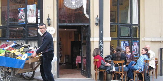 Menschen sitzen gemütlich vor einem Cafe, ein Mann steht an einem Obststand
