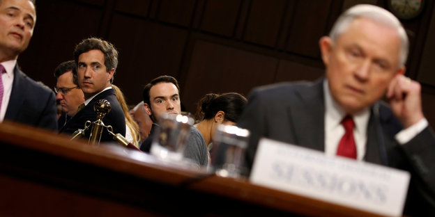 Jeff Sessions sitzt am Tisch und fasst sich ans Ohr, während ihn Journalisten im Hintergrund anschauen