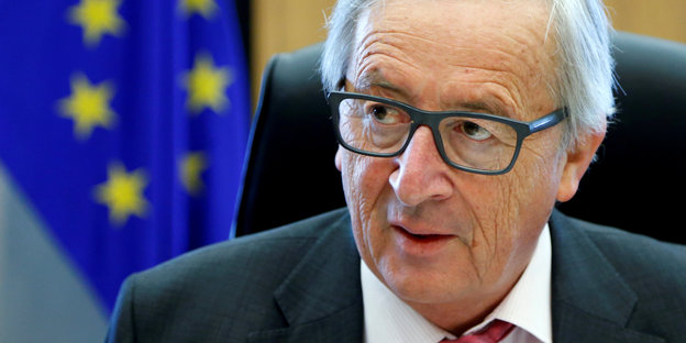 Juncker schaut durch seine Brille, im Hintergrund eine EU-Flagge