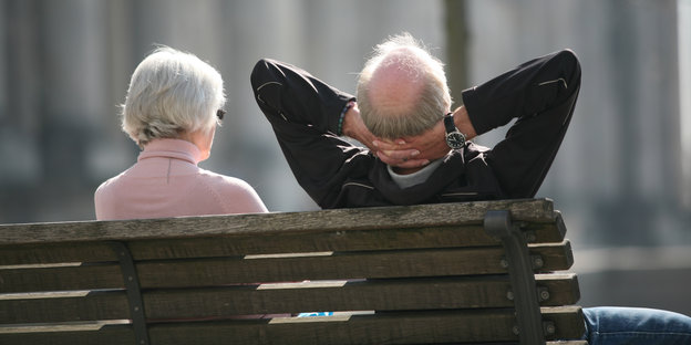 Zwei ältere Menschen sitzen nebeneinander auf einer Bank, der Mann hat die Arme hinter dem Kopf verschränkt.