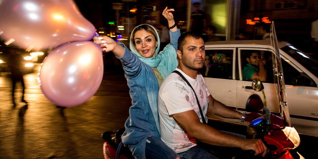 Eine Iranerin sitzt mit zwei Luftballons in der Hand hinten auf einem Motorroller, den ein Iraner fährt
