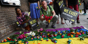 Mensche sitzen vor einer Regebogenfahne, auf der Blumen liegen