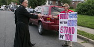 Ein Priester streckt den Arm einem Mann mit Plakat zum Handschlag aus, auf dem steht: "Good guys with guns stand by you"