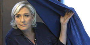 Marine Le Pen steht neben einem blauen Vorhang und zieht ihn halb über sich