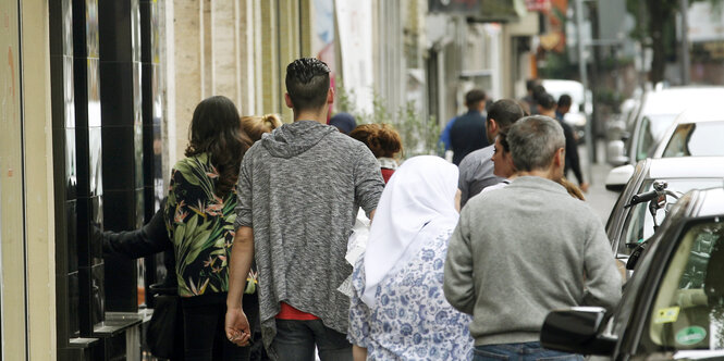 Menschen laufen zwischen Häuserfronten und geparkten Autos auf einem Gehweg