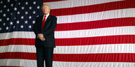 Ein Mann im Anzug steht vor einer riesengroßen US-Flagge